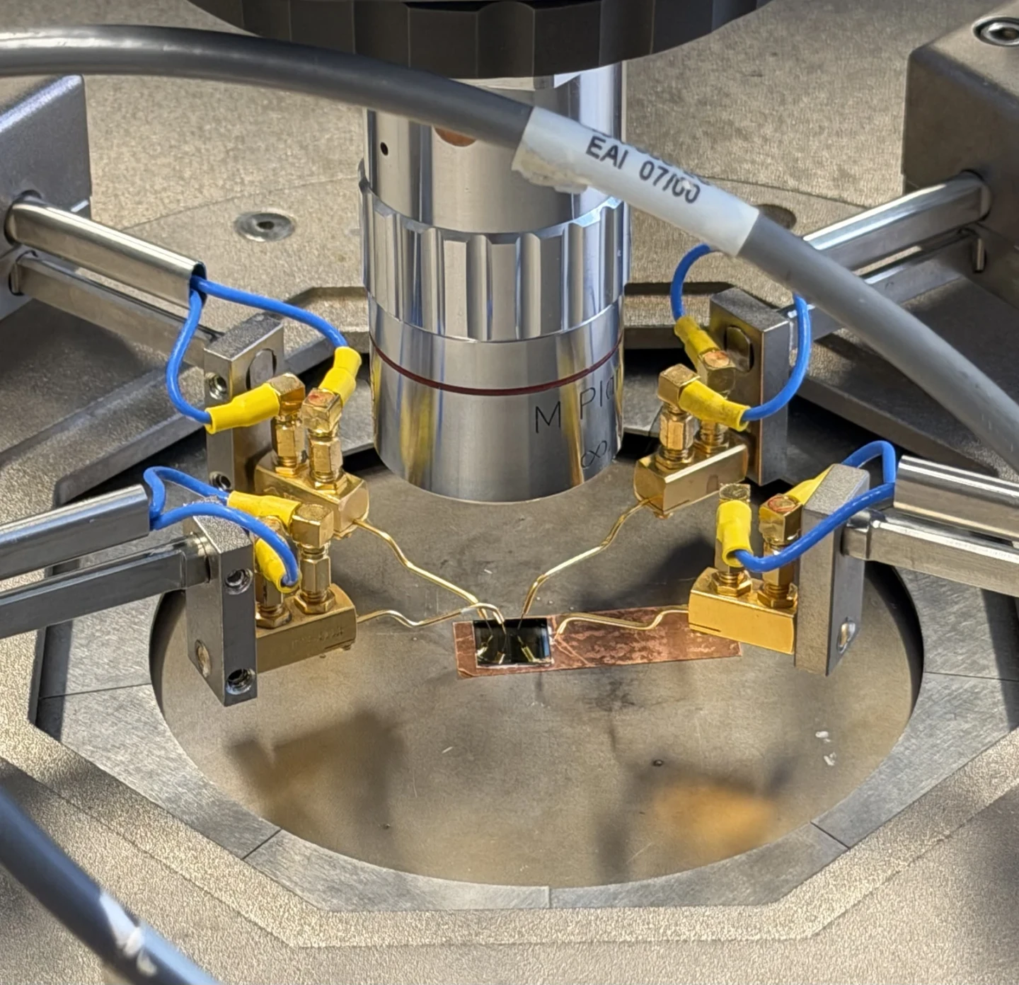 Four probe arms with gold probes and blue cables converge on a small chip on copper tape under a microscope objective.