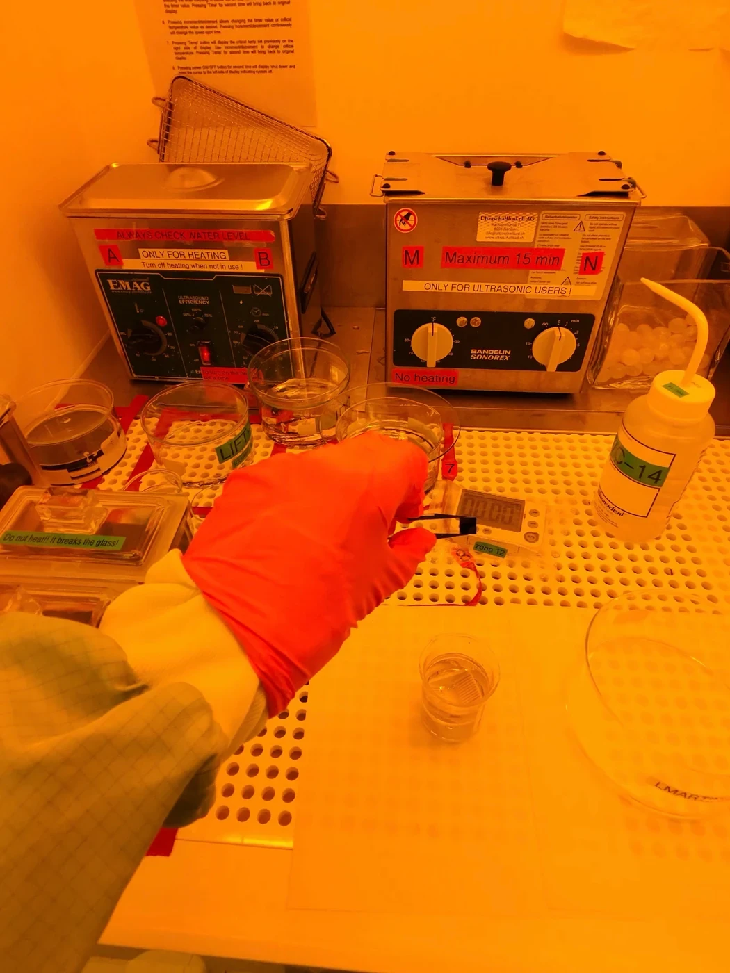 Under yellow cleanroom light, a gloved hand holds a small sample in tweezers over a wet bench with two ultrasonic baths and glass dishes.